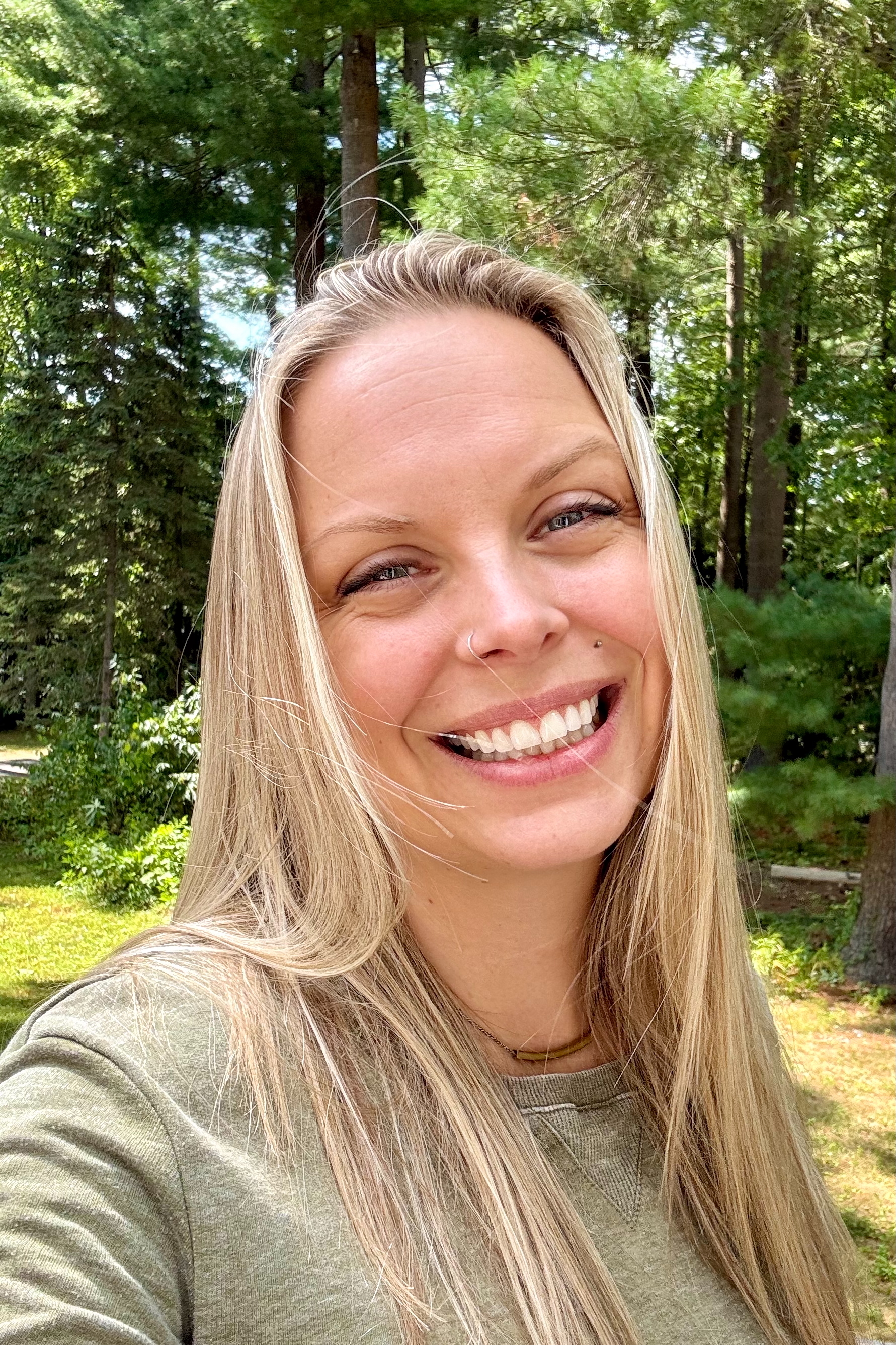 Lisa, a white woman with blonde hair, smiles at the camera with a background of green trees.